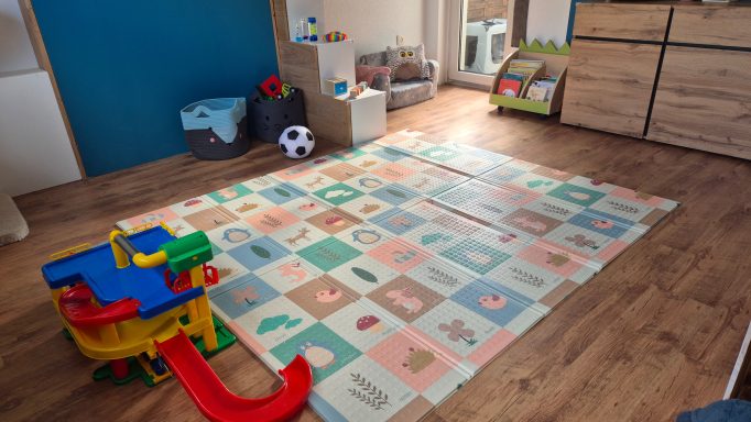 Bunte Spielmatte in einem Spielzimmer mit Spielzeug und Regalen im Hintergrund.