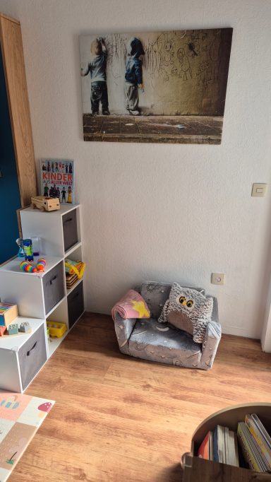 Spielzimmer mit einem kleinen Sofa, Spielzeugkisten und einem Wandbild von zwei Kindern.